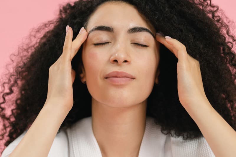 Woman with eyes closed resting her fingertips against her temples, a calm self-applied gesture like the finger drumming trick for tinnitus.