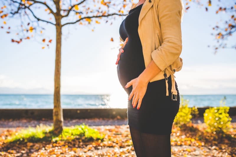 Pregnant woman in a beige jacket cradling her bump outdoors on a sunny autumn day by the sea.